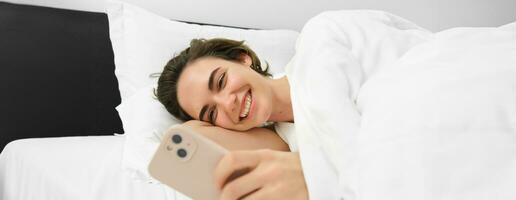 imagem do mulher deitado dentro cama com branco linho folhas, olhando às Smartphone e rindo, assistindo engraçado vídeo em Móvel telefone aplicativo foto