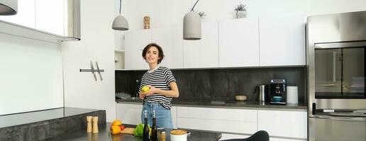 retrato do feliz, saudável jovem mulher, vegetariano fazer ela mesma salada, posando perto legumes em cozinha, cortar ingredientes para vegano refeição foto