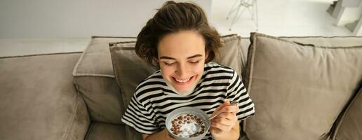 fechar acima do fofa jovem fêmea modelo, comendo cereais com leite, goza dela café da manhã em sofá dentro vivo sala, sorridente e olhando feliz foto