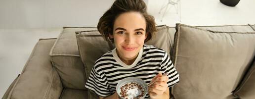 fechar acima do fofa jovem fêmea modelo, comendo cereais com leite, goza dela café da manhã em sofá dentro vivo sala, sorridente e olhando feliz foto