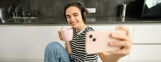 fechar acima retrato do mulher com Smartphone, levando selfie enquanto bebendo chá e ouvindo música dentro fones de ouvido, fazer fotos para dela social meios de comunicação aplicativo dentro a cozinha