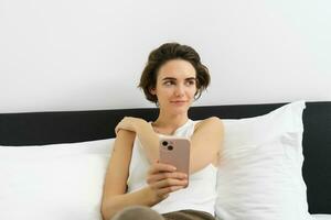 retrato do sorridente morena mulher em repouso dentro dela quarto, usando Móvel telefone e deitado dentro cama dentro dela casa roupa, Mensagens, enviando mensagem em Smartphone aplicativo foto