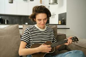 retrato do alegre jovem mulher jogando dela cavaquinho, cantando e rindo, sentado dentro vivo quarto às casa foto