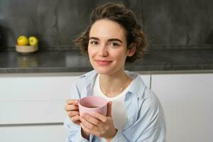 retrato do sorridente, lindo jovem mulher, bebendo café dentro cozinha, manhã Magia com cuppa chá, olhando ternamente foto