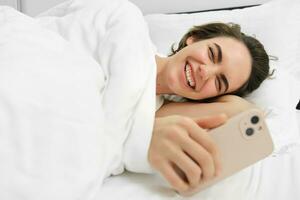 retrato do mulher assistindo engraçado videos dentro dela cama, olhando às Smartphone e rindo, usando Móvel telefone social meios de comunicação aplicativo antes indo para dormir foto