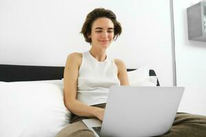retrato do sorridente moderno mulher, sentado em dela cama com computador portátil, olhando feliz, trabalhando a partir de lar, verificação dela e-mails em computador, navegando conectados lojas foto