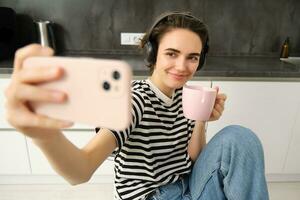 à moda fêmea modelo dentro fones de ouvido, leva selfie com copo do chá dentro a cozinha, postagem em social meios de comunicação aplicativo, usando Móvel telefone Câmera para faço conteúdo para dela blog foto