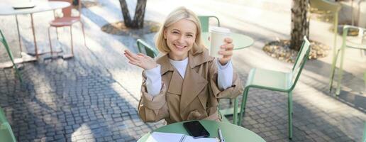 Comida e beber conceito. jovem feliz mulher sentado dentro cafeteria, desfrutando brilhante dia ao ar livre, levantando copo do café e sorridente, bebendo chai foto