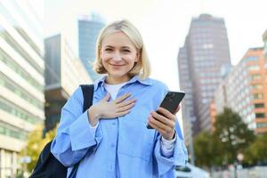 imagem do jovem profissional, escritório Gerente mulher com mochila e Smartphone, posando em ruas do ocupado cidade, segurando mão em peito, sorridente e olhando animado às Câmera foto