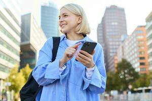 fechar acima retrato do à moda jovem Loiras mulher, em pé em rua, verificação dela Móvel telefone, usando Smartphone aplicativo para pegue por aí cidade, olhando às conectados mapa para instruções foto