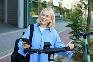 retrato do sorridente loiro mulher, Faculdade aluna usando Smartphone aplicativo para Varredura qr código em verde elétrico lambreta em rua, alugando uma passeio foto