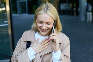 fechar acima retrato do sorridente, rindo jovem mulher, falando em Móvel telefone, em pé ao ar livre, caminhando em rua com Smartphone e conversando foto