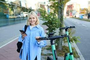 retrato do sorridente jovem mulher desbloqueia lambreta em rua, usando Móvel telefone para renda isto com Smartphone aplicativo, equitação para universidade foto