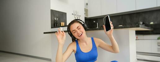 alegre morena menina faz ginástica às lar, desgasta sem fio fones de ouvido e danças, detém Smartphone, faz exercite-se dentro de casa em chão, desgasta azul roupa ativa foto