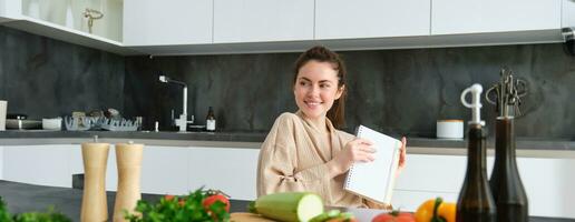 retrato do mulher escrevendo baixa Lista do mantimentos, fazer notas dentro receita, sentado dentro cozinha perto vegetais, preparando jantar cardápio foto