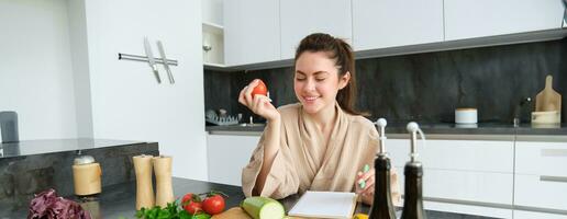 retrato do lindo jovem mulher dentro a cozinha, escrevendo baixa cozinhando receita, sentado perto cortar borda com legumes e fazer mercearia lista, criando saudável vegetariano cardápio para dela família foto