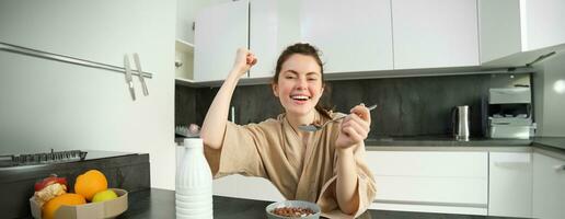 retrato do entusiasmado jovem mulher comendo cereais com leite, olhando animado e feliz, sentado perto cozinha bancada e tendo café da manhã, levantando mão acima dentro triunfo foto