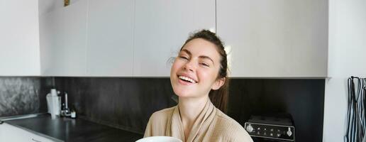 estilo de vida conceito. retrato do feliz morena mulher dentro roupão de banho, bebendo café dentro a cozinha, tendo manhã cuppa e sorridente foto