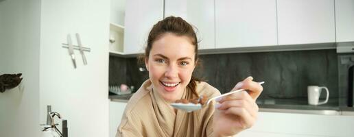 retrato do feliz jovem mulher inclina-se em cozinha bancada e comendo cereais, tem leite e tigela dentro frente do dela, tendo dela café da manhã, vestindo roupão de banho foto