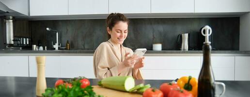 retrato do mulher dentro roupão de banho sentado dentro cozinha com Smartphone, cozinhando jantar, assistindo receita em social meios de comunicação, vídeo tutorial quão para preparar refeição foto