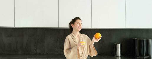 retrato do jovem e saudável mulher começa dela dia com frutas, comendo fresco laranja, bebendo suco dentro cozinha, vestindo roupão de banho foto