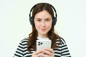 imagem do morena jovem mulher, sorridente, ouvindo para música dentro fones de ouvido, assistindo videos em Móvel telefone aplicativo, segurando Smartphone, em pé sobre branco fundo foto