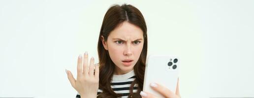 retrato do irritado menina encarando às dela Móvel telefone tela com frustrado face, branco fundo foto