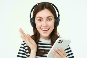 alegre, lindo jovem mulher usando Smartphone aplicativo, a comemorar, olhando feliz enquanto jogando com Móvel telefone, ouvindo música dentro sem fio fones de ouvido foto