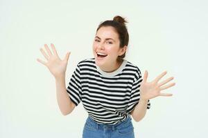 imagem do feliz, lindo jovem mulher faz jazz mãos, surpresa gesto, posando dentro casual camiseta e jeans sobre branco fundo foto