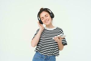 jovem mulher com Smartphone ouvindo para música, dançando para dela favorito música dentro fones de ouvido, posando contra branco fundo foto