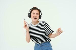 retrato do à moda jovem mulher dançando dentro sem fio fones de ouvido, usando fones de ouvido e Móvel telefone música aplicativo para ouço para favorito músicas, posando sobre branco fundo foto