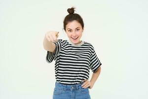 imagem do confiante, feliz jovem mulher dentro casual roupas, apontando dedo às Câmera, rindo e sorridente, em pé sobre branco fundo foto