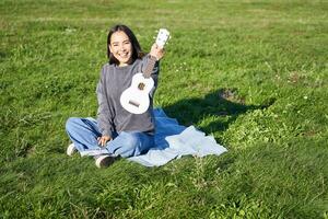 música e instrumentos. retrato do fofa ásia menina mostra dela branco cavaquinho, tocam dentro parque enquanto sentado relaxado em cobertor, desfrutando ensolarado dia foto