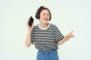 menina com Smartphone dançando, ouvindo para música dentro sem fio fones de ouvido, usando Móvel telefone transmissão aplicativo para apreciar favorito músicas, isolado sobre branco fundo foto