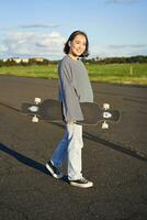 vertical tiro do despreocupado ásia menina com Longboard. jovem mulher skatista segurando cruzador em dela ombros e caminhando em estrada, skate foto