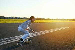 lado Visão do lindo ásia menina em skate, equitação dela cruzador para a Sol em a esvaziar estrada. feliz jovem skatista desfrutando ensolarado dia em dela patim foto