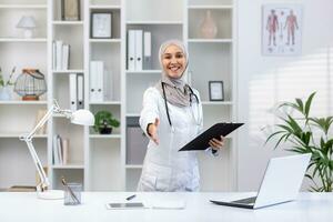 retrato do feliz muçulmano fêmea médico dentro hijab em pé às mesa dentro hospital escritório, vestindo branco casaco, pasta dentro mãos, estendendo mão dentro saudações, sorridente e olhando às Câmera. foto