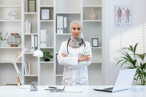 retrato do sério confiante bem sucedido fêmea médico dentro hijab dentro branco médico casaco olhando às Câmera com cruzado braços, clínica trabalhador trabalhando dentro escritório médico sala. foto