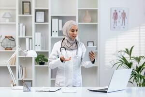 jovem com experiência fêmea médico dentro hijab usando tábua computador para vídeo chamar, fêmea clínica trabalhador consulta pacientes remotamente on-line, sorridente contente dentro clínica escritório. foto