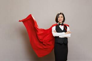 confiante sorridente garçonete vestindo Super heroi vermelho capa, mostrando força e poder. refeições serviço alegre mulher trabalhador dentro uniforme e Supermulher capa em pé com guardada mãos retrato foto