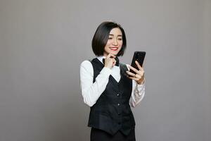 feliz ásia cafeteria garçonete lendo cliente positivo comentários em Smartphone. alegre sorridente recepcionista dentro profissional uniforme verificação cliente satisfação enquanto usando Móvel telefone foto