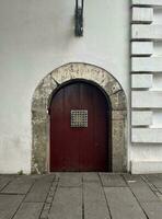 medieval estilizado vermelho porta com volta arco e pequeno quadrado metal rede janela isolado em branco muro. ao ar livre europeu velho era estilizado exterior arquitetura fotografia isolado em vertical Razão imagem. foto