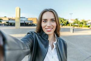 bonita jovem mulher com Loiras cabelo e azul olhos sorridente enquanto levando uma selfie em a rua foto