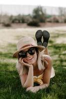 foto vertical de uma linda jovem com um chapéu de praia e óculos escuros deitada na grama