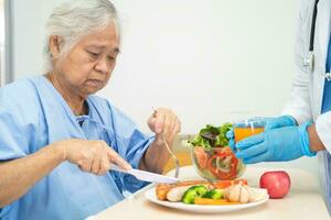 ásia Senior mulher paciente comendo salmão bife café da manhã com vegetal saudável Comida enquanto sentado e com fome em cama dentro hospital. foto