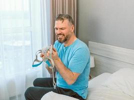 homem maduro e feliz com problemas respiratórios crônicos, considera usar a máquina de cpap sentada na cama no quarto. saúde, cpap, conceito de ronco foto