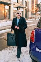 mulher elegante sênior em elegante casaco preto e bolsa de mão, caminhando de seu carro para uma reunião de negócios. negócio, estilo, conceito anti-idade