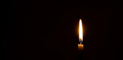 uma solteiro queimando vela chama ou luz brilhando em uma pequeno laranja vela em Preto ou Sombrio fundo em mesa dentro Igreja para Natal, velório ou memorial serviço com cópia de espaço foto
