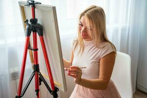 jovem mulher artista com paleta e escova sentado e pintura cenário em tela de pintura perto janela. arte e criatividade conceito. Alto qualidade foto