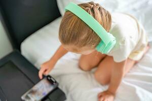 fofa pequeno menina dentro verde crianças fones de ouvido é usando uma Smartphone, tentando para encontrar a certo música para ouvindo. Alto qualidade foto